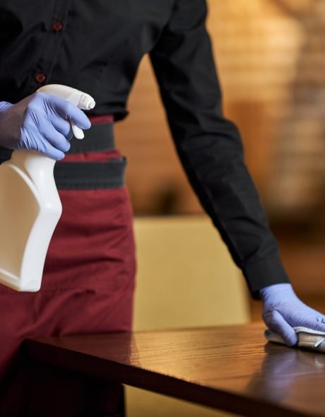 Cropped photo of restaurant employee wearing disposable gloves and holding spray bottle while disinfecting a table